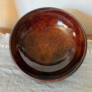 May include: A handmade, glazed ceramic bowl with a rich, earthy brown color. The bowl has a speckled texture and a glossy finish, with a dark brown rim. The interior reflects light, highlighting the depth of the glaze.