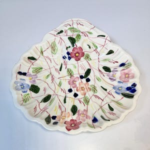 May include: A white, shell-shaped ceramic serving dish with a scalloped edge. The dish is decorated with pink, purple, and blue flowers, green leaves, and thin red vines. The floral design covers the entire surface of the dish.
