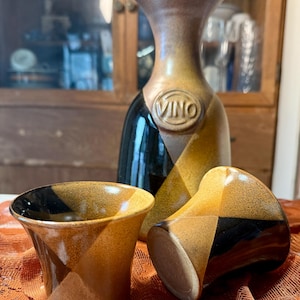 May include: A ceramic wine decanter and two matching cups. The decanter is brown and black with the word "VINO" embossed on the front. The cups and decanter have a geometric design.