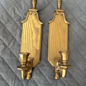 May include: A pair of gold-colored wall sconces featuring a vertical wooden plaque with a visible grain texture, topped with a decorative finial. Each sconce includes a protruding candle holder cup attached to the lower section of the plaque.