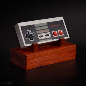 May include: A wooden display stand for a classic Nintendo controller. The stand is made of dark wood and features a rectangular base with a matching support for the controller. The controller is white and grey with red buttons and the Nintendo logo.