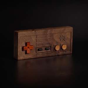 May include: A wooden video game controller with a dark brown finish. The controller features a red directional pad, two black buttons, and two wooden buttons. The design is reminiscent of a classic gaming console controller.