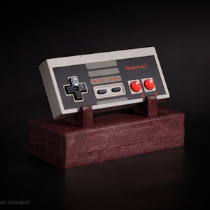 Op de afbeelding: Een vintage Nintendo-controller, grijs en zwart, rust op een donkerhouten standaard. De controller heeft rode knoppen en het Nintendo-logo. De standaard is donkerroodbruin.
