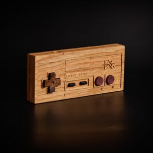 Puede incluir: Un mando de juego de madera con un diseño clásico. El mando presenta una cruceta direccional, dos botones rojos y una forma rectangular. La madera tiene una veta natural.