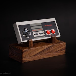 Peut inclure: Une manette Nintendo posée sur un présentoir en bois. La manette est grise et noire avec des boutons rouges et le logo Nintendo. Le présentoir est un bloc rectangulaire brun foncé avec des supports en bois.