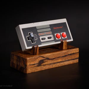 May include: A Nintendo controller, with red buttons and a directional pad, rests on a wooden stand. The controller is gray and black, with the Nintendo logo in red. The stand is made of dark wood with a visible grain.