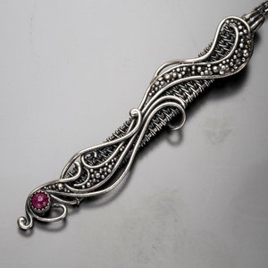 Puede incluir: Un collar con un colgante de plata, con un diseño delicado e intrincado que presenta un patrón de alambre tejido y una sola piedra preciosa roja rubí.