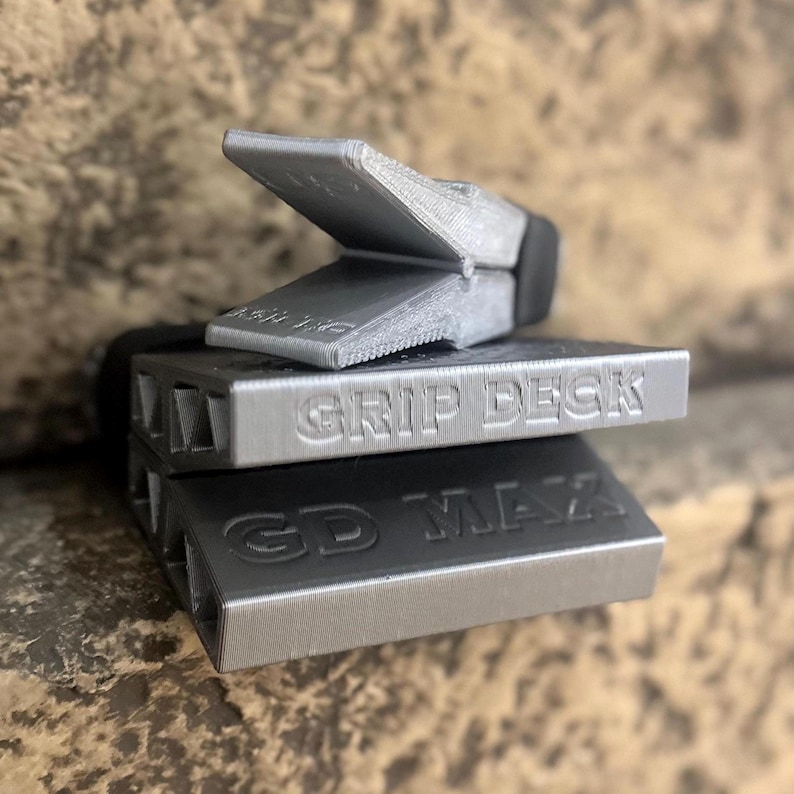 Puede incluir: Tres objetos rectangulares de color plateado apilados uno encima del otro. El objeto superior tiene una pieza articulada y en &aacute;ngulo. El objeto del medio est&aacute; etiquetado "GRIP DECK" y el inferior "GD MAX".