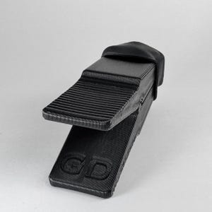 Puede incluir: Una cuña de nivelación negra impresa en 3D con una superficie estriada. La cuña tiene forma rectangular con un borde biselado y una pequeña parte superior redondeada. Las letras "GD" están grabadas en la base.