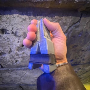 Puede incluir: Una herramienta gris y negra con una parte superior acanalada y una banda negra. La herramienta se sostiene en una mano contra un fondo de piedra texturizada. La herramienta parece estar hecha de pl&aacute;stico o un material similar.
