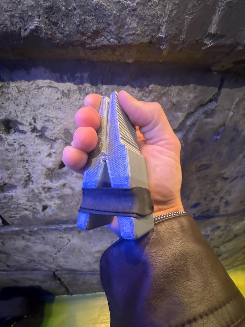 Puede incluir: Una herramienta gris y negra con una parte superior acanalada y una banda negra. La herramienta se sostiene en una mano contra un fondo de piedra texturizada. La herramienta parece estar hecha de pl&aacute;stico o un material similar.