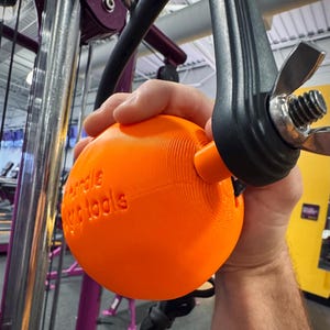 May include: An orange, spherical weight training handle with the text "Handle Magic Tools" embossed on its surface. The handle is attached to a black metal frame with a wing nut. The background shows gym equipment.