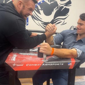 Puede incluir: Dos hombres compitiendo en pulso en una mesa con "COMBAT ARM SPORTS" impreso. La mesa es roja y negra. Un mural de dos brazos musculosos est&aacute; en el fondo. Un hombre lleva una sudadera con capucha negra, el otro una camisa azul.