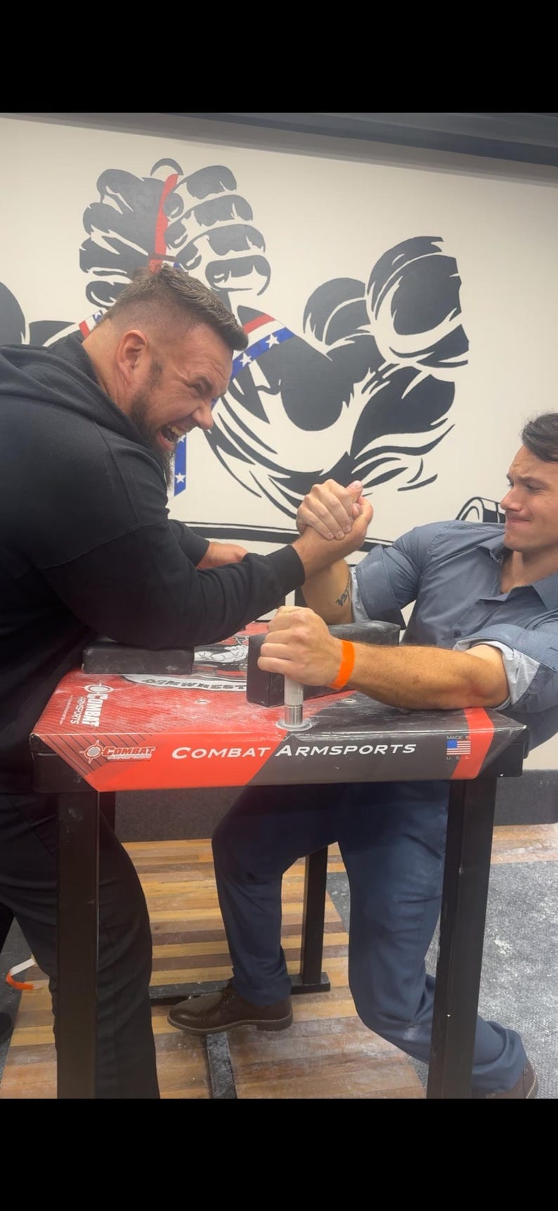 Puede incluir: Dos hombres compitiendo en pulso en una mesa con "COMBAT ARM SPORTS" impreso. La mesa es roja y negra. Un mural de dos brazos musculosos est&aacute; en el fondo. Un hombre lleva una sudadera con capucha negra, el otro una camisa azul.