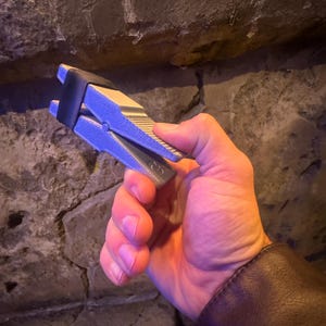 Puede incluir: Un objeto met&aacute;lico plateado y negro con un dise&ntilde;o de resorte, sostenido en una mano. El objeto tiene una superficie texturizada y una banda de goma negra. El fondo es una pared de piedra rugosa.