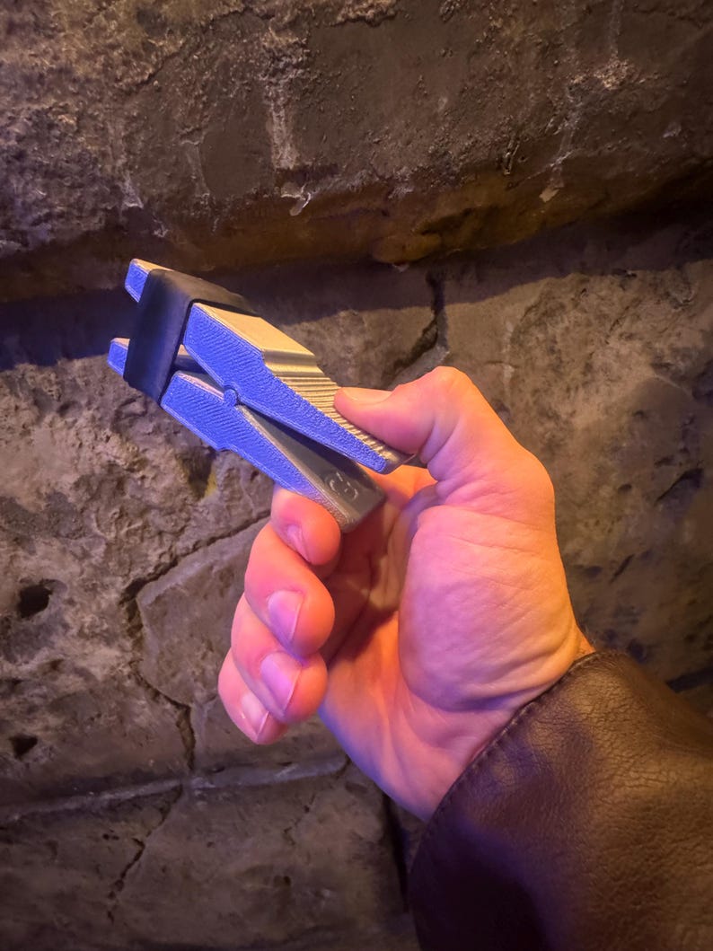 Puede incluir: Un objeto met&aacute;lico plateado y negro con un dise&ntilde;o de resorte, sostenido en una mano. El objeto tiene una superficie texturizada y una banda de goma negra. El fondo es una pared de piedra rugosa.