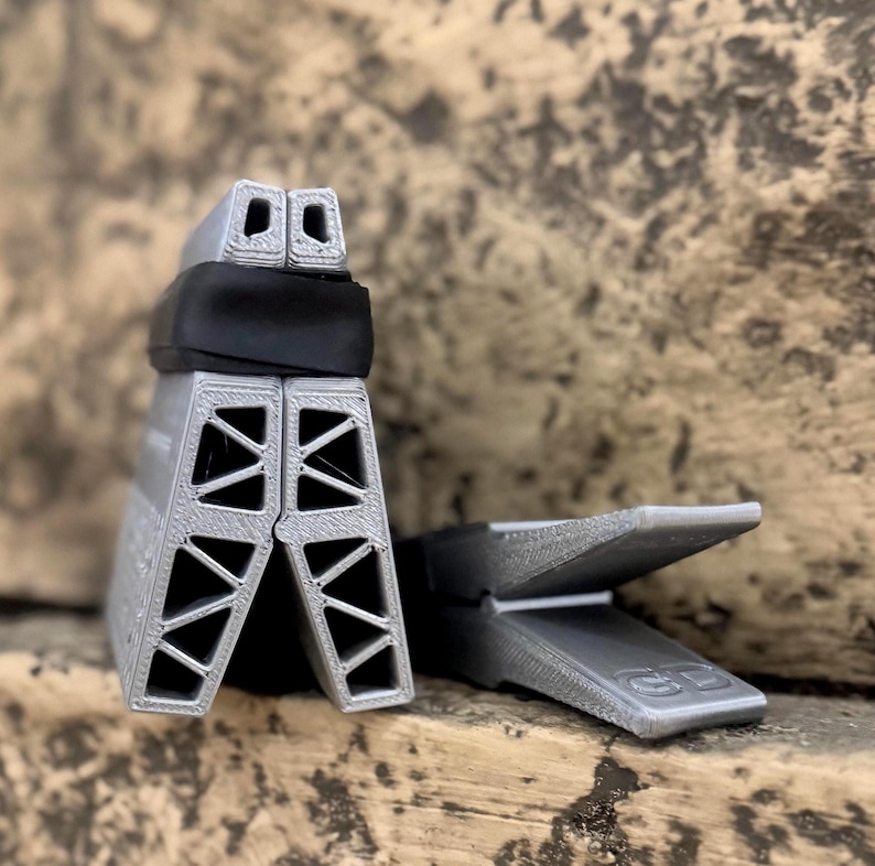 Puede incluir: Dos objetos grises de forma geom&eacute;trica. Uno est&aacute; en posici&oacute;n vertical, con una banda negra alrededor de la parte superior. El otro est&aacute; tumbado. Ambos tienen cortes triangulares. Los objetos parecen ser de material impreso en 3D.