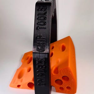 May include: A black grip tool with the text "GRIPDECK GRIP TOOLS" is holding two orange cheese-shaped objects. The cheese objects have multiple holes and a textured surface. The grip tool has two silver metal rods.