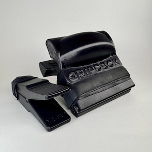 Może przedstawiać: Dwa czarne produkty Gripdeck są wyświetlane na białym tle. Większy przedmiot ma wytłoczony napis "GRIPDECK" z boku. Mniejszy przedmiot ma kształt klina z żebrowaną powierzchnią. Oba przedmioty wydają się być wykonane z twardego tworzywa sztucznego.