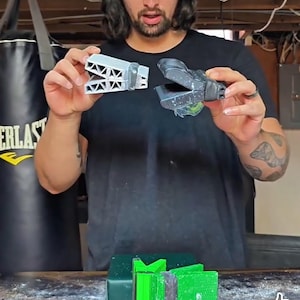 Puede incluir: Una persona sostiene dos herramientas de agarre GripDeck. Una herramienta es gris con un dise&ntilde;o triangular, y la otra es negra. Varias herramientas GripDeck de colores est&aacute;n sobre una mesa, incluyendo rojo, verde y gris.