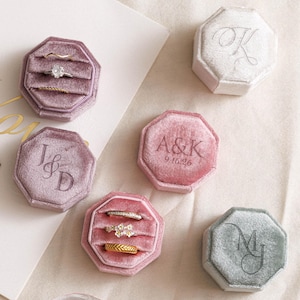May include: Multiple octagonal velvet ring boxes in shades of mauve, pink, and gray, some featuring engraved initials and dates. Two open boxes display various metal rings, while closed boxes show personalized monograms on their lids.