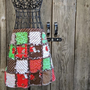 以下が含まれることがあります： 茶色、赤、白、緑の配色でパッチワークされたスカート。スカートは、ストライプ、水玉模様、クリスマスモチーフなど、さまざまなパターンで構成された正方形の布で作られています。スカートは黒い金属製のマネキンに飾られています。