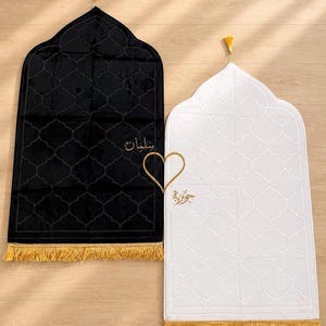 Puede incluir: Dos alfombras de oración rectangulares, una negra y otra blanca, con una parte superior arqueada y una borla dorada. Ambas alfombras tienen un patrón de cuatrifolio repetitivo, flecos dorados en la base y caligrafía árabe bordada.