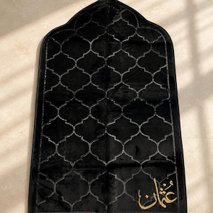 May include: A black prayer rug featuring a repeating arched lattice pattern across the surface. The top is pointed with a small gold tassel, and the bottom edge has gold fringe. The Arabic name Othman is embroidered in gold thread in the corner.