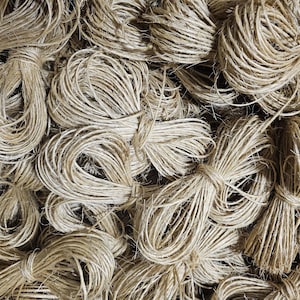 May include: A close-up of a pile of natural twine, made from jute or hemp, in a variety of sizes and lengths. The twine is a light brown color and is tightly wound into balls.
