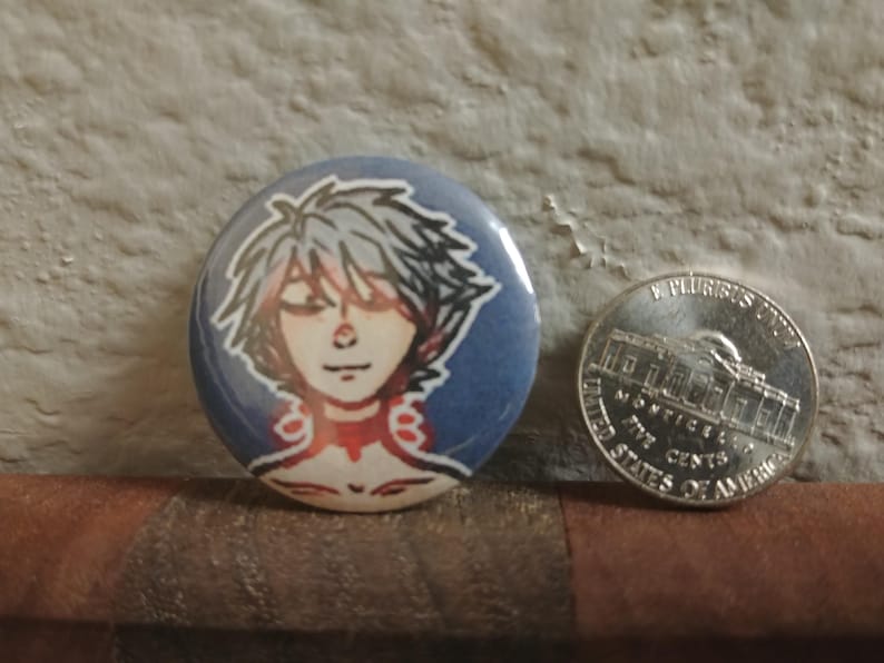 May include: A round button with a blue background and a white character with silver hair. The character has red and white accents around their neck.