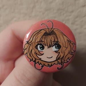 May include: A pink button with a cartoon drawing of a girl with brown hair and green eyes. The girl is smiling and has a light pink blush on her cheeks.