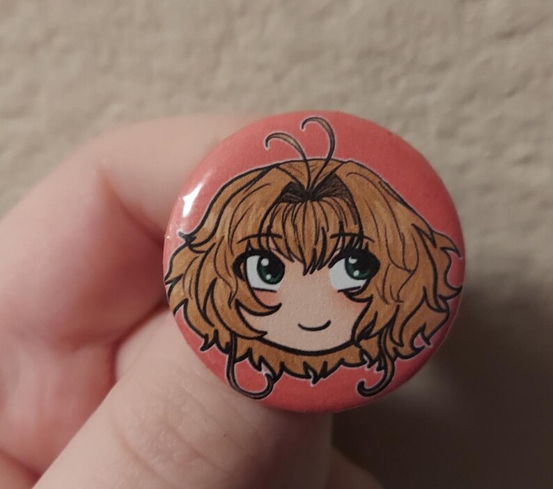 May include: A pink button with a cartoon drawing of a girl with brown hair and green eyes. The girl is smiling and has a light pink blush on her cheeks.