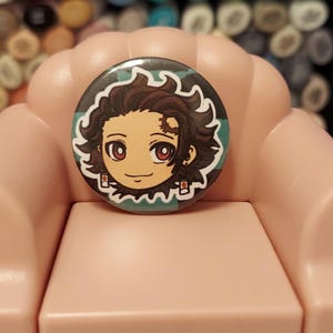 May include: A round button featuring a cartoon illustration of a young person with brown hair and green eyes. The person is wearing a green and white checked shirt. The button is displayed on a pink armchair.