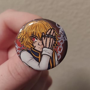 May include: A round button featuring a cartoon illustration of a character with blond hair, red eyes, and a chain covering one eye. The character is wearing a white shirt with a blue collar and a gold trim. The background is red with a chain pattern.