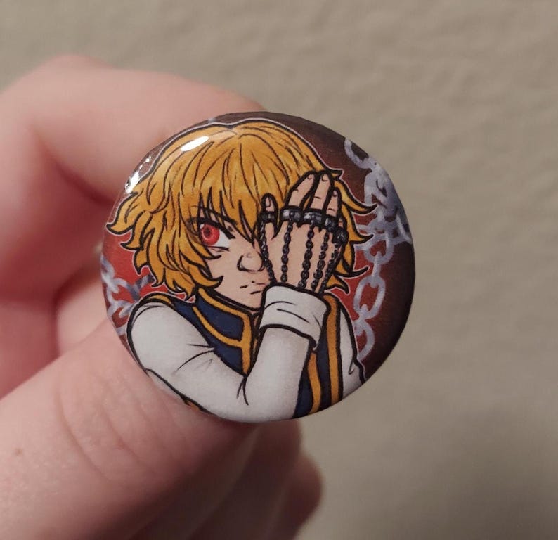 May include: A round button featuring a cartoon illustration of a character with blond hair, red eyes, and a chain covering one eye. The character is wearing a white shirt with a blue collar and a gold trim. The background is red with a chain pattern.