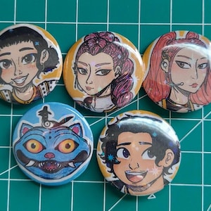 May include: Five round buttons with colorful illustrations. The buttons feature portraits of people and a blue cat-like creature. The portraits have various hair colors and styles. The buttons are on a green surface.