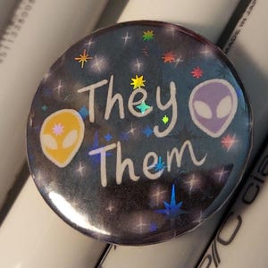 May include: A round button with a dark blue background featuring the words "They Them" in white script. The button has two alien head illustrations, one yellow and one purple, and holographic star accents.