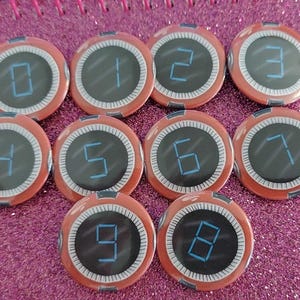 Puede incluir: Una colección de diez botones redondos, rojos y negros, cada uno mostrando un número diferente del 0 al 9 en una fuente de estilo digital. Los números son azules. Los botones están dispuestos sobre una superficie rosa brillante.