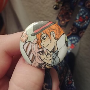 May include: A round button featuring a cartoon illustration of two people. The person on the left is wearing a pink and white striped dress and has brown hair. The person on the right is wearing a red and black hat and a white shirt. The background is green.
