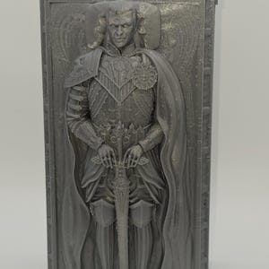 May include: A gray, rectangular relief sculpture of a man in armor holding a sword. The man is depicted in a regal pose, wearing a cape and ornate chest armor. The sculpture is framed within a rectangular border.