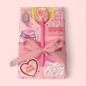 May include: A pink gift box tied with a ribbon, containing a lollipop, pen, stickers, and sticky notes. The stickers read "Hot Girls Read Books" and "Book Lover". A bookmark with the text "Reading is never a waste of time" is also visible.