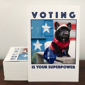 May include: A stack of white cards with the words "VOTING IS YOUR SUPERPOWER" printed on them. The cards feature a black French Bulldog dressed as a superhero, with a red and blue costume and a gold headband.