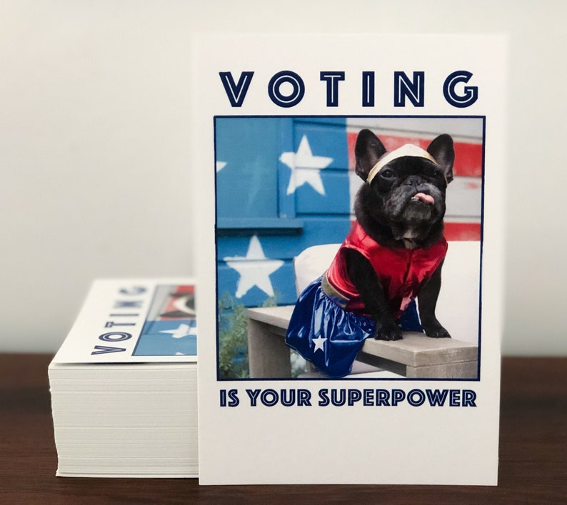 May include: A stack of white cards with the words "VOTING IS YOUR SUPERPOWER" printed on them. The cards feature a black French Bulldog dressed as a superhero, with a red and blue costume and a gold headband.
