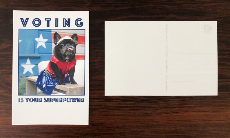 May include: A postcard featuring a black French Bulldog dressed in a superhero costume. The dog is wearing a red vest and blue skirt with a white star. The text "VOTING IS YOUR SUPERPOWER" is printed on the card.