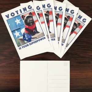 May include: A stack of white postcards with a patriotic theme. Each card features a black French bulldog dressed in a superhero costume, with the text "VOTING IS YOUR SUPERPOWER" in blue. The background includes a painted American flag.