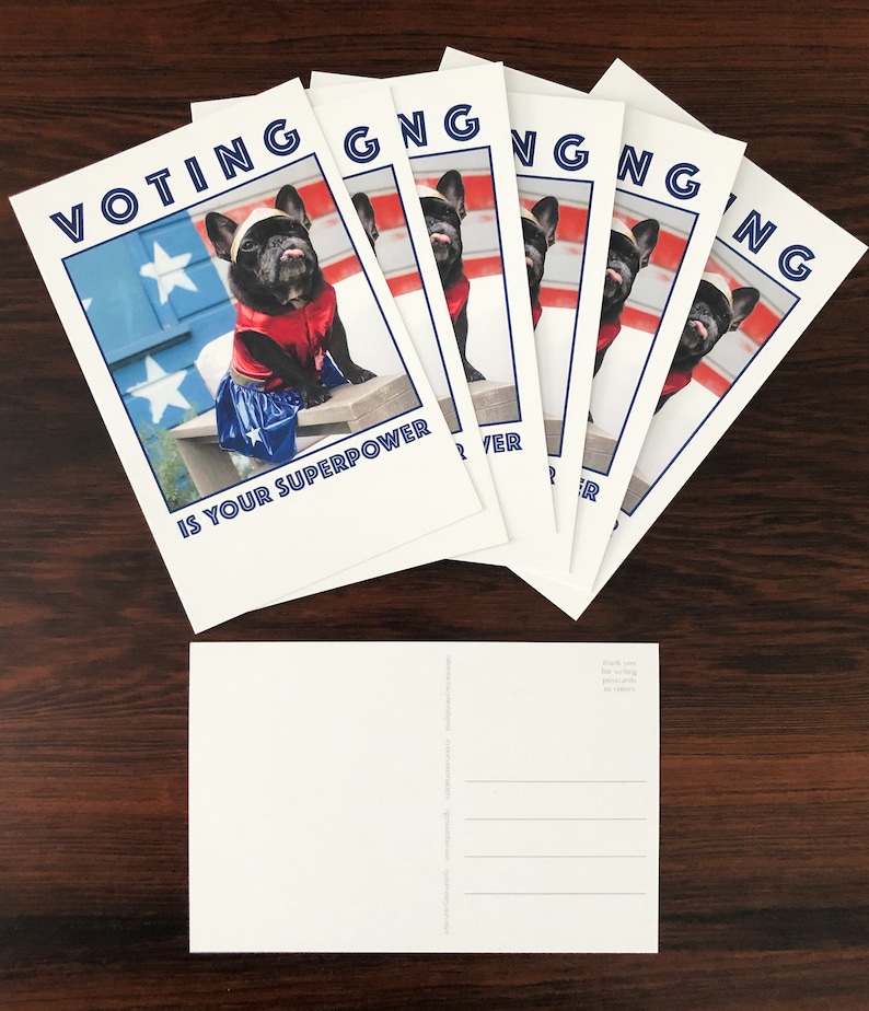 May include: A stack of white postcards with a patriotic theme. Each card features a black French bulldog dressed in a superhero costume, with the text "VOTING IS YOUR SUPERPOWER" in blue. The background includes a painted American flag.