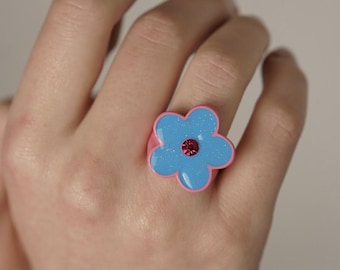 Op de afbeelding: Een roze bloemenring met een blauw centrum en een glinsterende afwerking. De ring wordt aan een vinger gedragen.