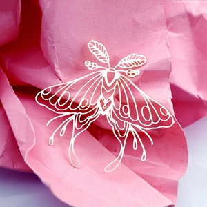 Puede incluir: Corte de papel blanco de una polilla con detalles intrincados, sobre un fondo de papel rosa arrugado.