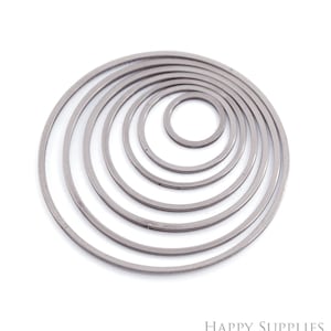 Puede incluir: Un conjunto de siete colgantes circulares de metal plateado de diferentes tamaños, dispuestos de forma concéntrica. El círculo más grande está en la parte inferior, el más pequeño en el centro. Estos colgantes son probablemente para la fabricación de joyas o artesanía.