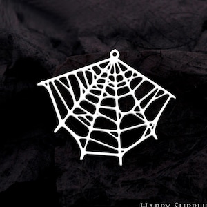 Puede incluir: Colgante blanco calado con forma de telaraña, con un pequeño lazo en la parte superior para colgarlo. El diseño de la telaraña presenta una serie de líneas interconectadas, creando un patrón detallado e intrincado. El colgante está sobre un fondo oscuro.